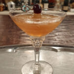 A light amber liquid is shown in a cut glass coupe glass. A beaded silver cocktail pick rests on top, holding a cherry.