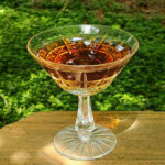 A cut glass coupe cocktail glass sits on a wooden plank surrounded by greenery. The liquid within is a clear, rich pinkish orange.