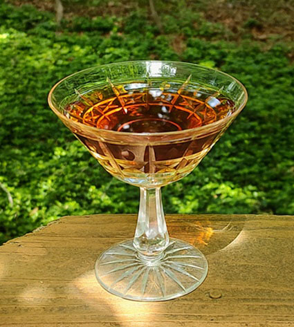 A cut glass coupe cocktail glass sits on a wooden plank surrounded by greenery. The liquid within is a clear, rich pinkish orange.