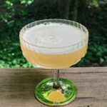 A scalloped glass coupe glass featuring a green base holds a freshly shaken, light yellow-orange cocktail.