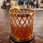The Sic Semper Tyrannis cocktail in a vintage gold lattice-patterned Imperial Glass rocks glass, deep amber-red in color and garnished with a lemon twist on a vintage sword cocktail pick, resting on a round coaster atop a wooden bar counter with bottles softly blurred in the background.