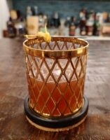 The Sic Semper Tyrannis cocktail in a vintage gold lattice-patterned Imperial Glass rocks glass, deep amber-red in color and garnished with a lemon twist on a vintage sword cocktail pick, resting on a round coaster atop a wooden bar counter with bottles softly blurred in the background.