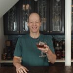 Josh Wulf holding an amber-colored Mooch cocktail in a coupe glass behind his home bar, with bottles, glassware, and Art Deco-style cabinets in the background.