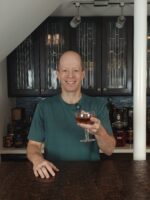 Josh Wulf holding an amber-colored Mooch cocktail in a coupe glass behind his home bar, with bottles, glassware, and Art Deco-style cabinets in the background.
