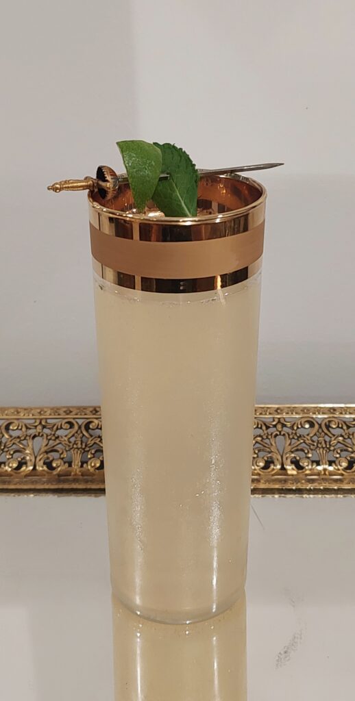 Mojito in a tall vintage glass with wide gold bands around the rim, set on a Hollywood-style vintage tray and garnished with lime and mint held by a vintage Toledo sword cocktail pick.