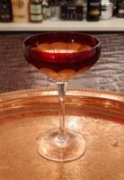 Widow’s Kiss cocktail served in a Waterford crystal coupe with a deep red bowl and clear crystal stem, resting on an ornate gold-toned tray with engraved floral patterns.