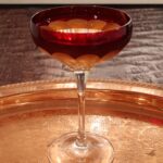 Widow’s Kiss cocktail served in a Waterford crystal coupe with a deep red bowl and clear crystal stem, resting on an ornate gold-toned tray with engraved floral patterns.