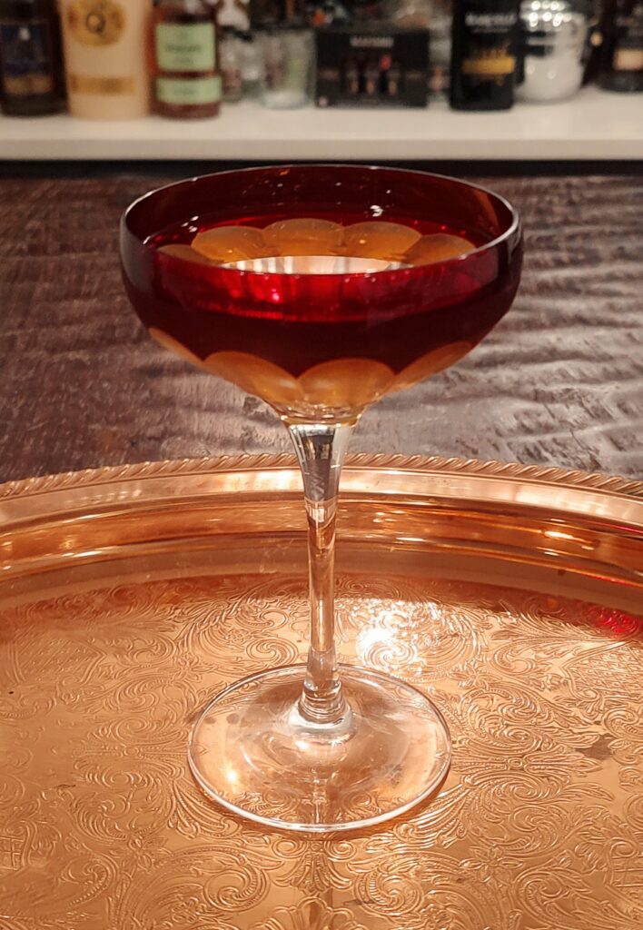 Widow’s Kiss cocktail served in a Waterford crystal coupe with a deep red bowl and clear crystal stem, resting on an ornate gold-toned tray with engraved floral patterns.